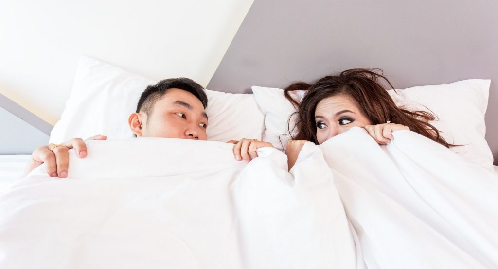 Asian couple in bed peeking out from under the covers
