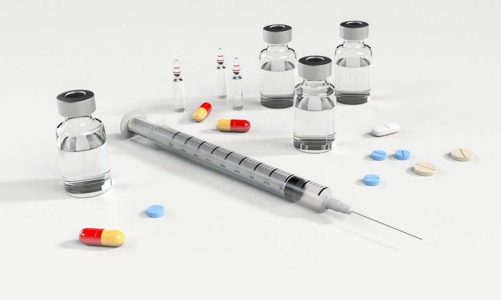 all types of vaccines - vials of vaccine, red and yellow pills, blue pills, white pills and brown pills laying next to vials and needle with vaccines